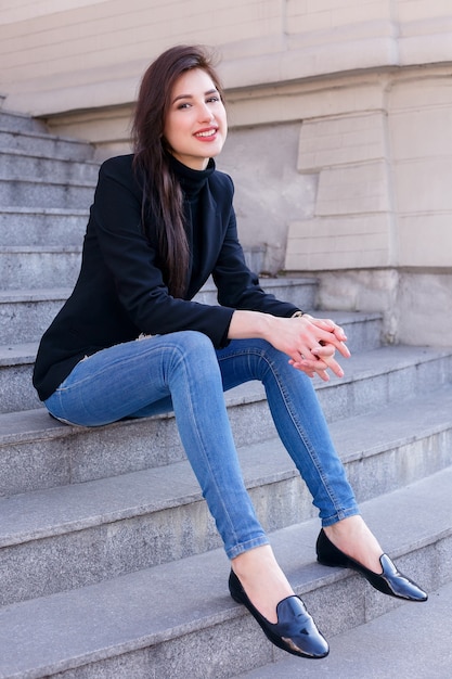 Chica en una chaqueta negra y jeans azul sentado en pasos.