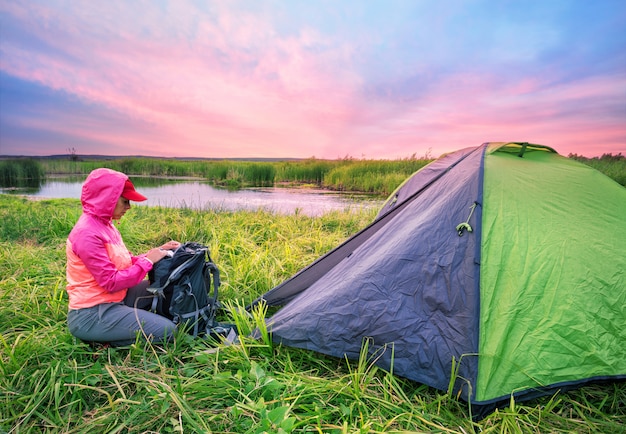 Chica de chaqueta rosa abre su mochila cerca de la carpa en el río ba