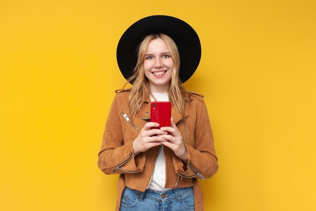 Una chica con una chaqueta y un sombrero de otoño sostiene un teléfono con un fondo amarillo aislado