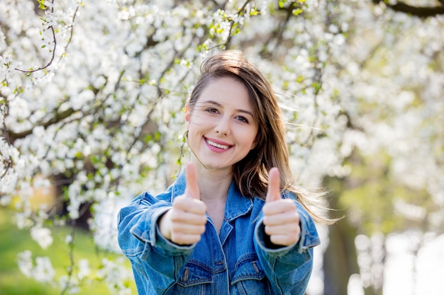 La chica con una chaqueta vaquera se encuentra cerca de un árbol en flor y muestra el signo de la mano Ok