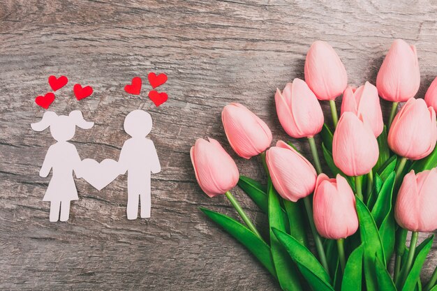 La chica y el chico están cortados de papel, sobre un fondo de madera, contra el fondo de un ramo de tulipanes rosados.