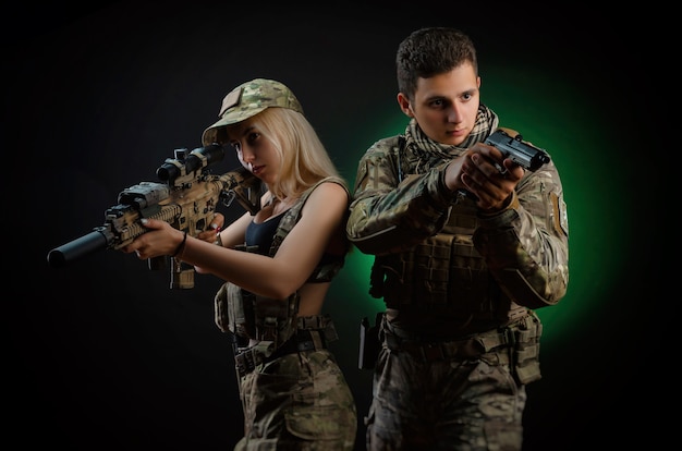 Una chica y un chico con un mono militar posan con una pistola de airsoft sobre un fondo oscuro