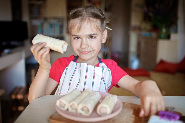 Chica cocinando salchichas enrolladas en hojaldre para el desayuno, el pequeño chef disfruta de un sabroso aperitivo