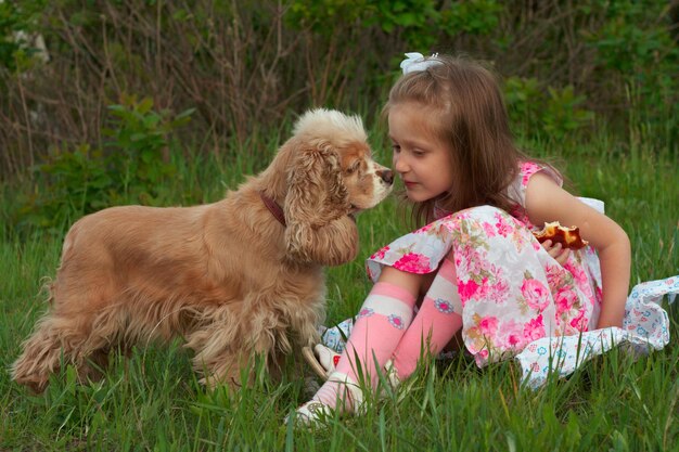 Chica comiendo un bollo sentada en un césped y su perro mirándolo de cerca