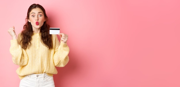 Foto chica compradora emocionada diciendo que sí haciendo puño y mirando asombrada mostrando tarjeta de crédito de plástico y rea