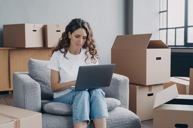 Chica comprando en línea una casa nueva en una computadora portátil sentada entre cajas de tarjetas Comercio electrónico de reubicación