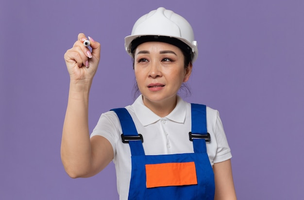 Chica constructora asiática joven complacida con casco de seguridad blanco sosteniendo y mirando el marcador