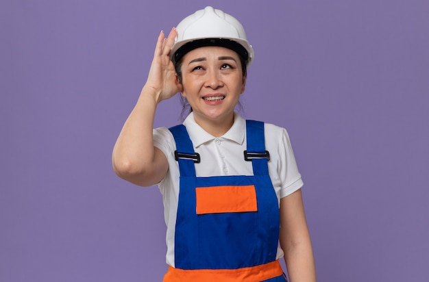 Chica constructora asiática joven disgustada poniendo la mano en su casco de seguridad blanco y mirando hacia arriba