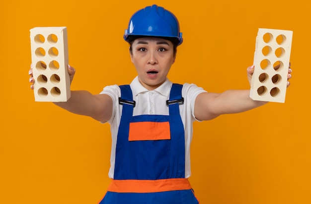 Foto chica constructora asiática joven sorprendida con casco de seguridad azul sosteniendo ladrillos