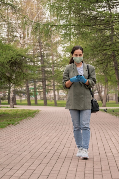 Chica contemporánea en máscara y ropa casual elegante en smartphone