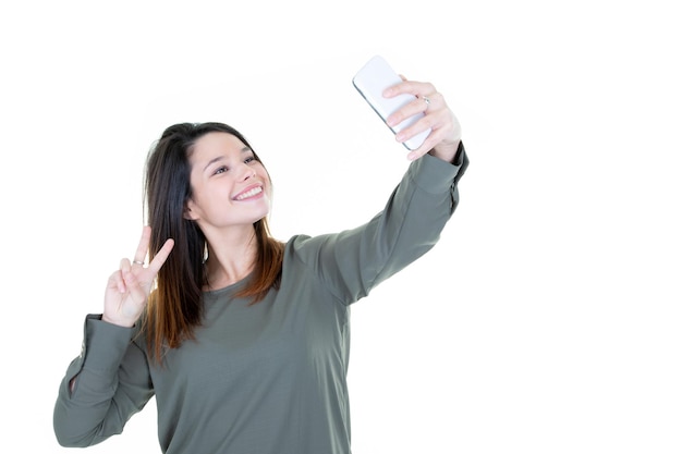 Chica cool de moda tomando un retrato de selfie en un teléfono inteligente haciendo el signo de la paz sobre fondo blanco