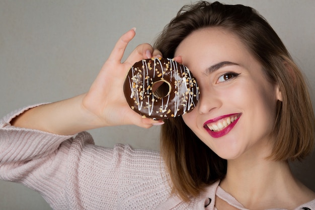 Chica coqueta alegre con maquillaje natural con donut sobre un fondo gris. Espacio para texto