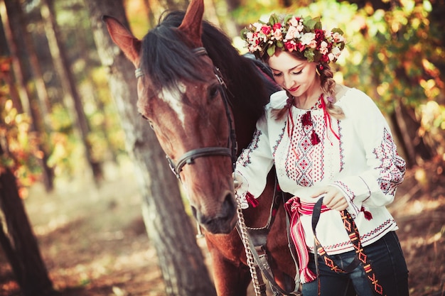 Chica en una corona y camisa bordada con un caballo en el bosque