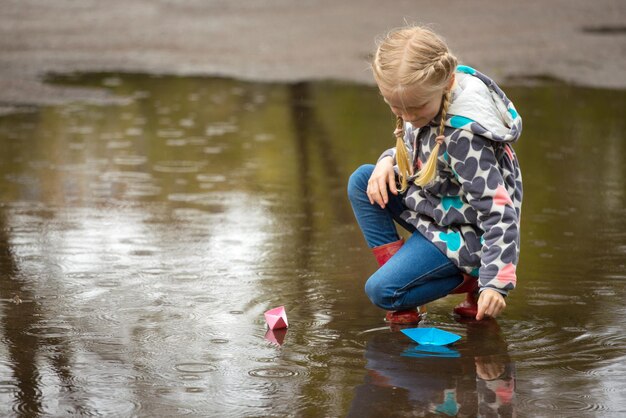 Chica corre el barco de papel rosa en un charco bajo la lluvia