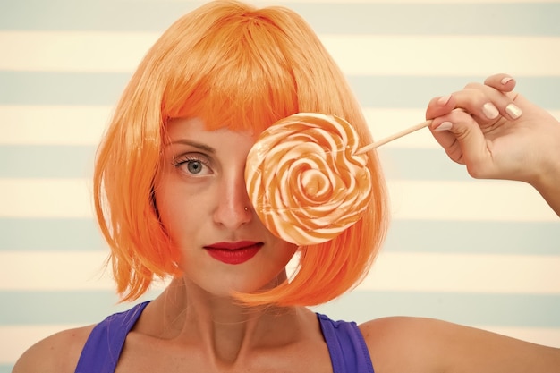 La chica cosplay con una mirada loca sostiene una piruleta naranja. La chica se ve infantil y sexy con labios rojos.