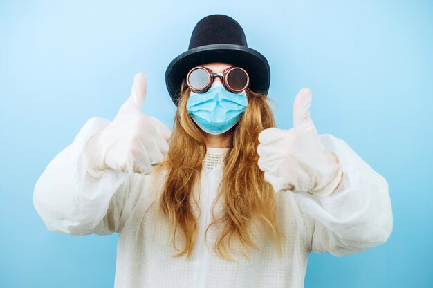 Foto chica creativa en una túnica blanca, máscara médica, gafas y un sombrero sobre un fondo azul. autoaislamiento durante la cuarentena de coronavirus. epidemia covid-2019.