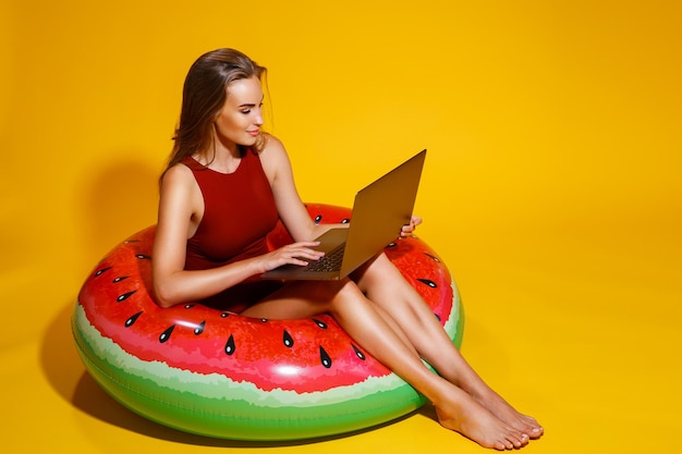 Chica de cuerpo entero en bikini sentada en un anillo de sandía inflable usa una computadora portátil en un viaje de fondo amarillo