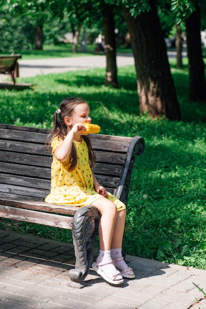 Una chica de cuerpo entero con un vestido amarillo está sentada en el borde de un banco en un parque de verano y ...