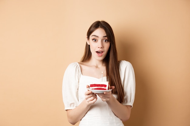 Chica de cumpleaños emocionada mirando sorprendido, sosteniendo pastel con vela, haciendo deseo de b-día, de pie sobre fondo beige.