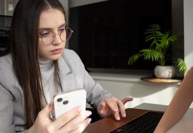 Chica dama de negocios sosteniendo un teléfono inteligente en sus manos y mirando una computadora portátil