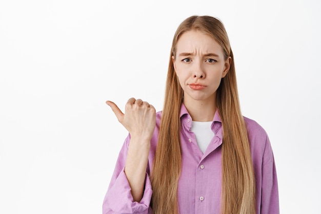 Foto una chica decepcionada parece escéptica apuntando con el dedo a la izquierda en algo malo y poco interesante producto de pie contra el fondo blanco sonríe y frunce el ceño