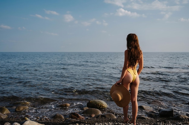 Chica delgada con un botín sexy con un sombrero de paja en traje de baño en la playa junto al mar