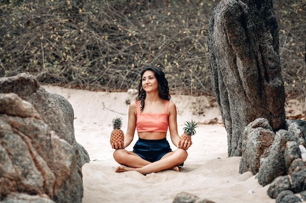 Chica delgada feliz haciendo yoga en la playa y sosteniendo dos piñas.