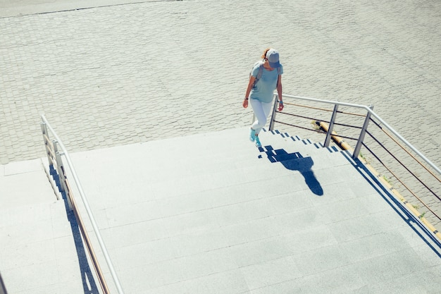 Chica delgada con gorra y vaqueros subiendo escaleras en la ciudad en el verano