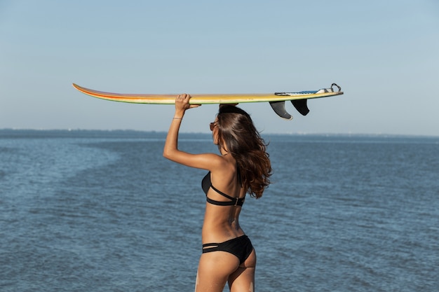 Chica delgada de pelo oscuro en traje de baño negro sostiene una tabla de surf amarilla sobre su cabeza cerca del mar en un día soleado