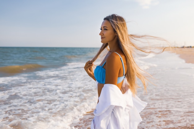 Una chica delgada en un traje de baño azul suave y una camisa camina por la playa de arena cerca del mar azul