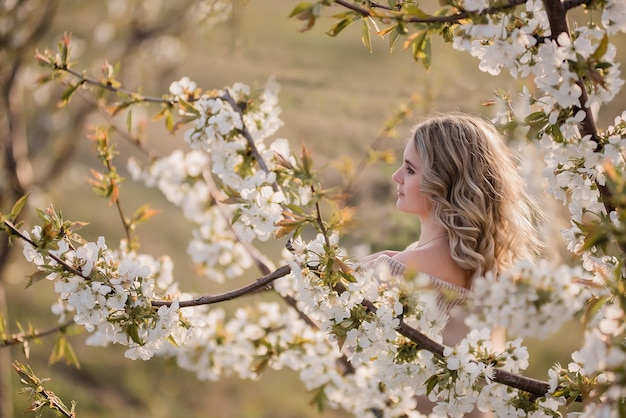 Chica delicada y soñadora con cabello rubio rizado en jardines blancos florecientes al atardecer