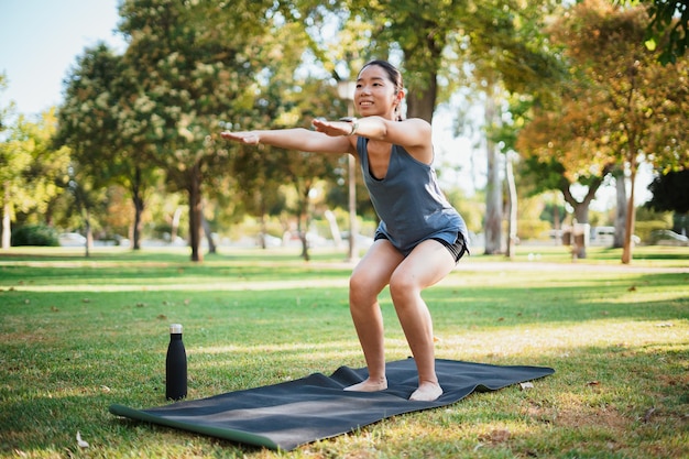 Chica deportiva asiática haciendo ejercicio al aire libre en el parque. Está haciendo ejercicios en cuclillas en una colchoneta.