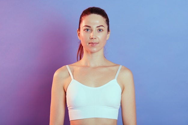 Chica deportiva atlética en camiseta blanca sin mangas, posando después de ejercicios de entrenamiento y mirando directamente a la cámara, tiene cola de caballo, de pie contra la pared de color.