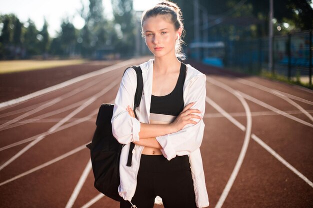 Chica deportiva en auriculares inalámbricos con mochila en el hombro mirando cuidadosamente a la cámara en la cinta de correr del estadio