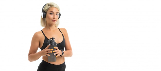 Una chica deportiva con cabello rubio y manicura brillante en auriculares, temas deportivos negros y polainas sostiene una botella de agua deportiva.