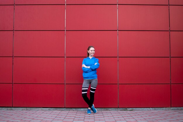 Chica deportiva se levanta contra una pared roja en ropa deportiva, fitness mujer posando en una calle de la ciudad, espacio de copia