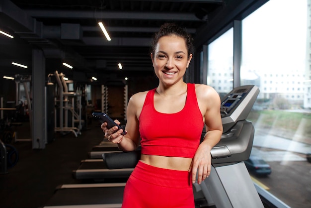 Chica deportiva en ropa deportiva roja usa teléfono inteligente en el gimnasio y sonríe mujer delgada sostiene el teléfono