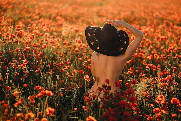 Chica desnuda por detrás con un sombrero negro en un campo de amapolas al atardecer