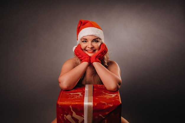 Foto chica desnuda con un sombrero de navidad se sienta con un regalo en un fondo negro