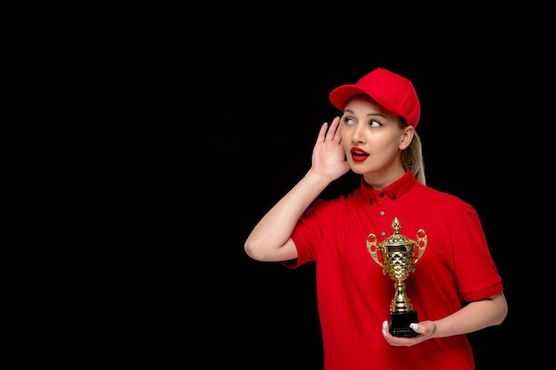 Chica de día de camisa roja sosteniendo un trofeo escuchando a escondidas en una gorra roja con camisa y lápiz labial brillante
