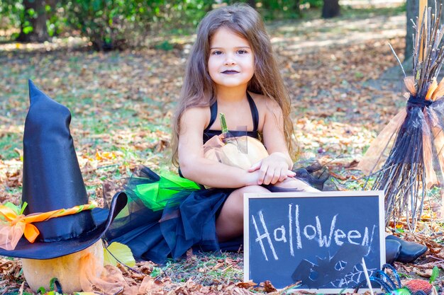 Chica en un disfraz de bruja para la fiesta de Halloween. Cartel con la inscripción: Halloween. Niña sentada en la hierba