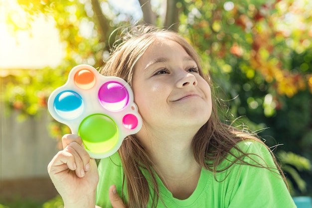 Una chica divertida se esconde detrás de un juguete. Retrato de niña con un juguete popit moderno. Pop it colorido y brillante juguete simple hoyuelo. antiestrés juguete sensorial fidget push simple hoyuelo