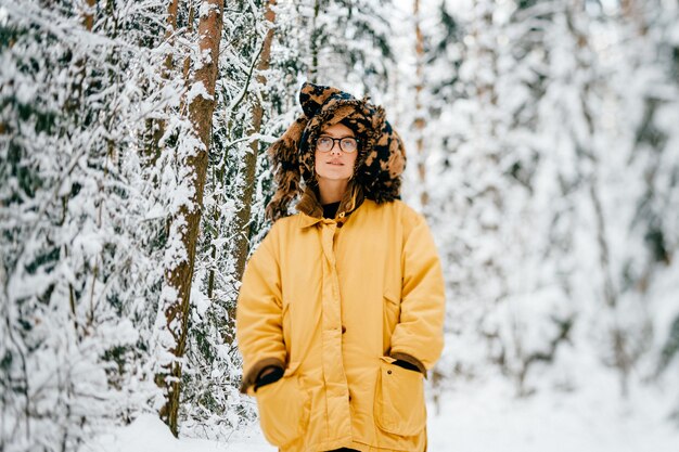 Chica divertida joven inconformista en chaqueta amarilla, gafas y turbante de la bufanda posando en el bosque de nieve