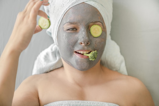 Chica divertida con máscara cosmética hecha de arcilla gris y trozos de verduras en la cara, su envoltura de cabello