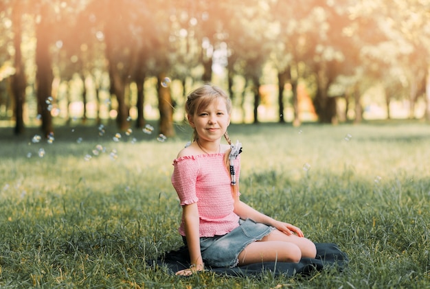 Chica con dos coletas se sienta en la hierba en el verano cerca del río. Niño sonriente en el jardín de verano