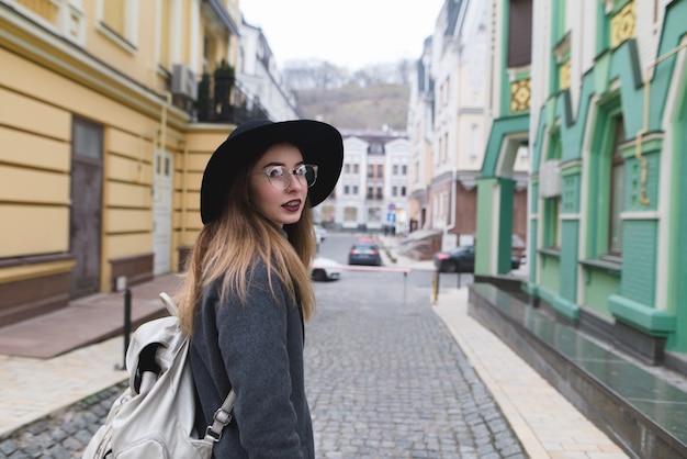 Chica elegante con un abrigo y sombrero camina por el casco antiguo y mira a la cámara. hermosa mujer viaja