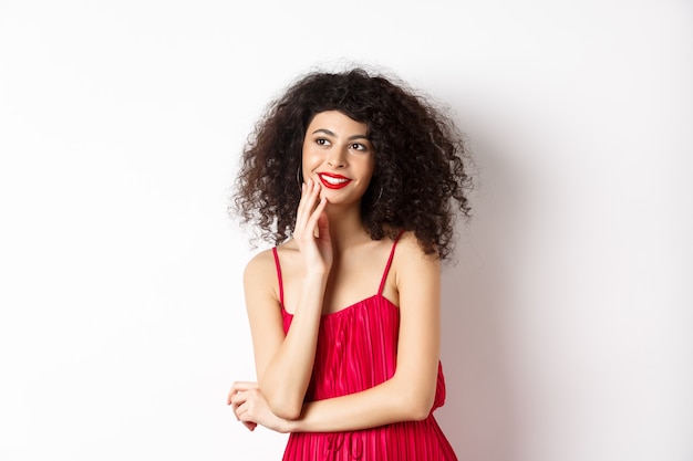 Chica elegante de ensueño con pelo rizado, vestido rojo y maquillaje, mirando a la izquierda y sonriendo al logo, de pie sobre fondo blanco.