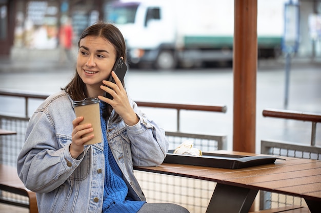 Chica elegante en estilo casual habla por teléfono con café en la mano y está esperando a alguien.