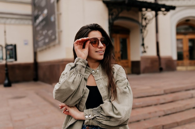 Chica elegante y fresca con chaqueta de mezclilla mira hacia otro lado Atractiva mujer bronceada con chaqueta beige y gafas de sol camina al aire libre y se divierte