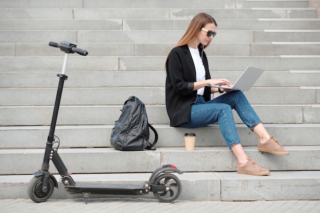 Chica elegante con laptop navegando en la red en la escalera con su mochila y bebida cerca mientras se toma un descanso después de caminar en scooter eléctrico